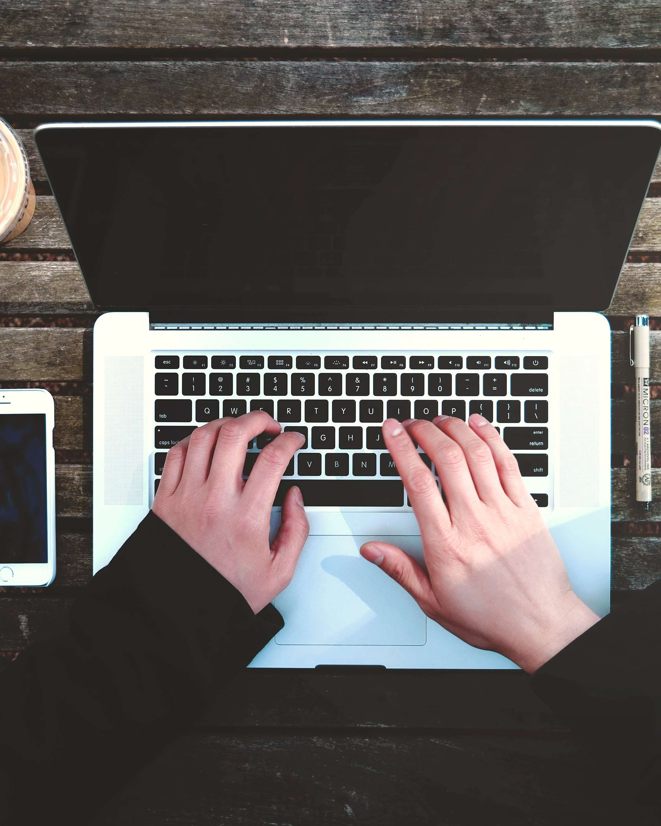 Hands typing on a laptop on a dark wooden table, a phone and a set square beside it