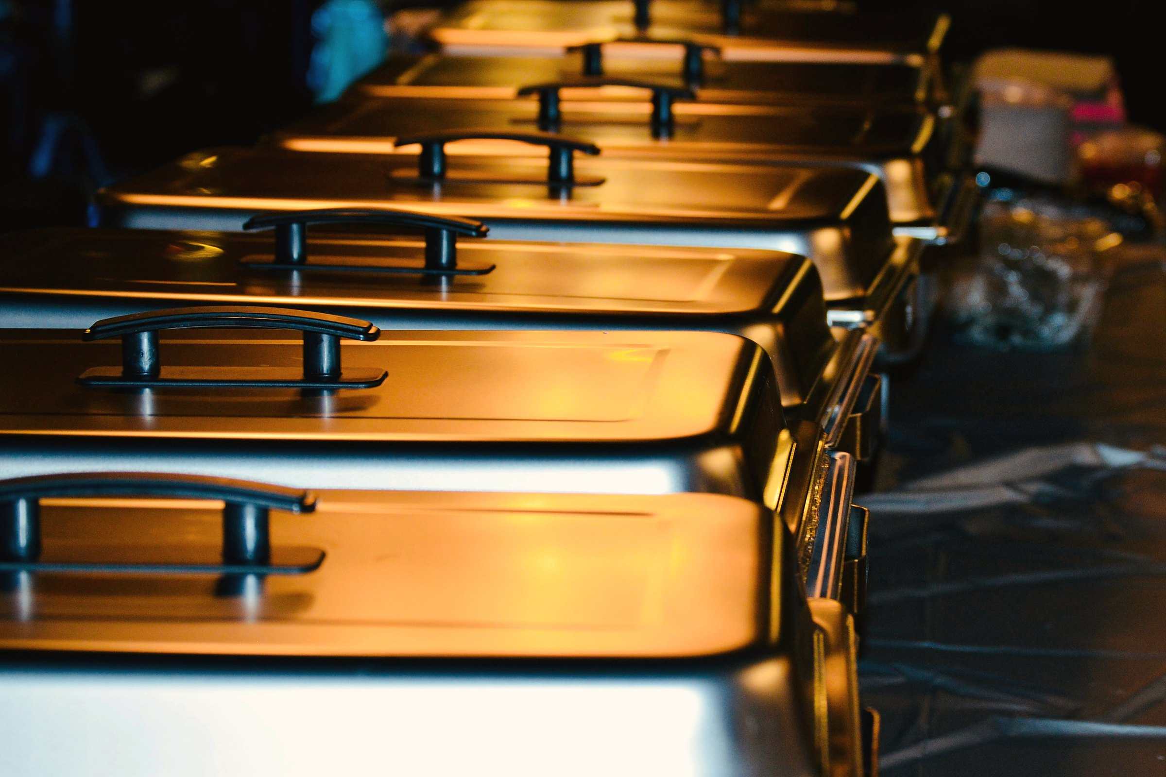 Rows of covered steel chafing dishes glowing under warm light at a catered event