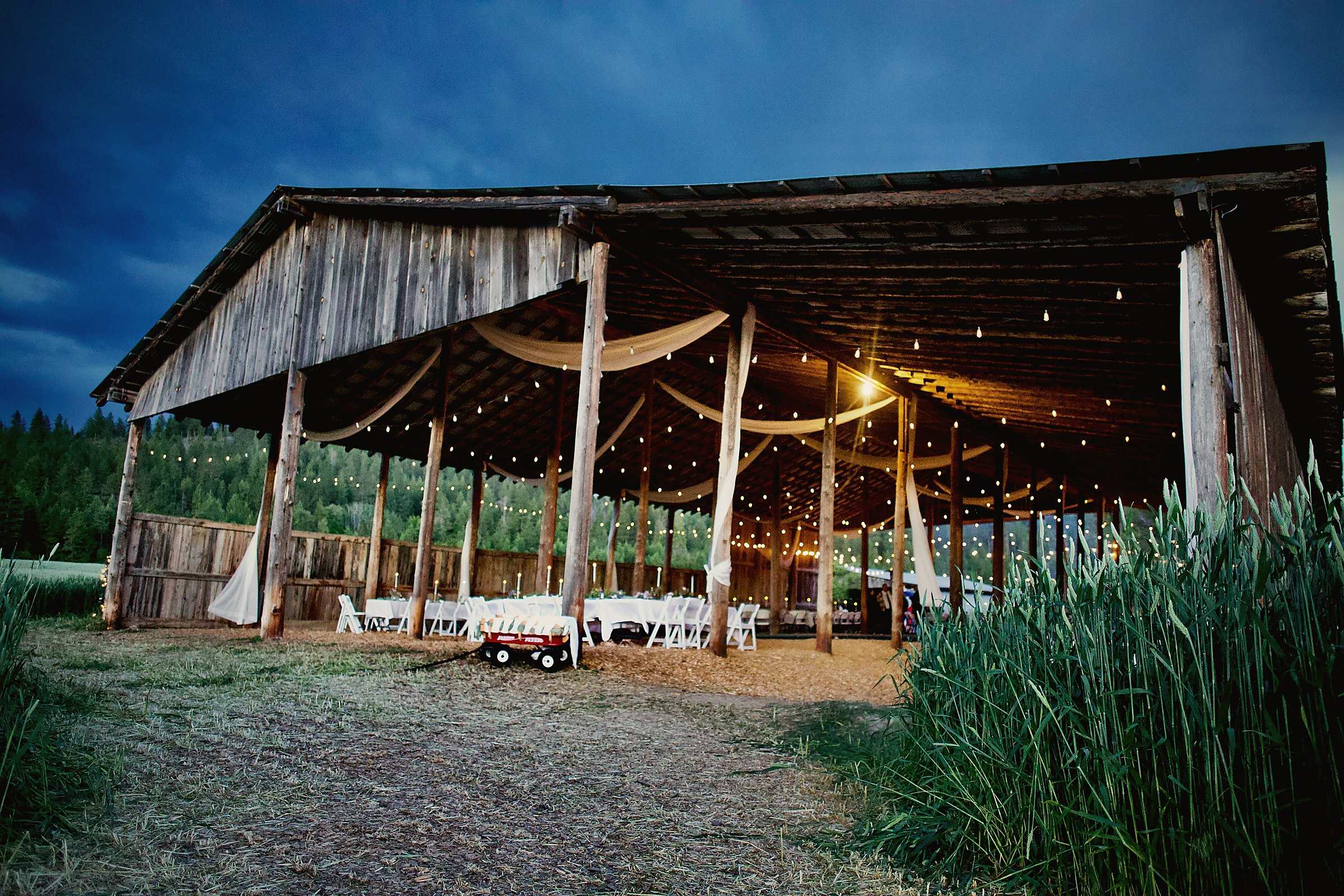 An open wooden barn hung with glowing string lights at twilight, tables set inside