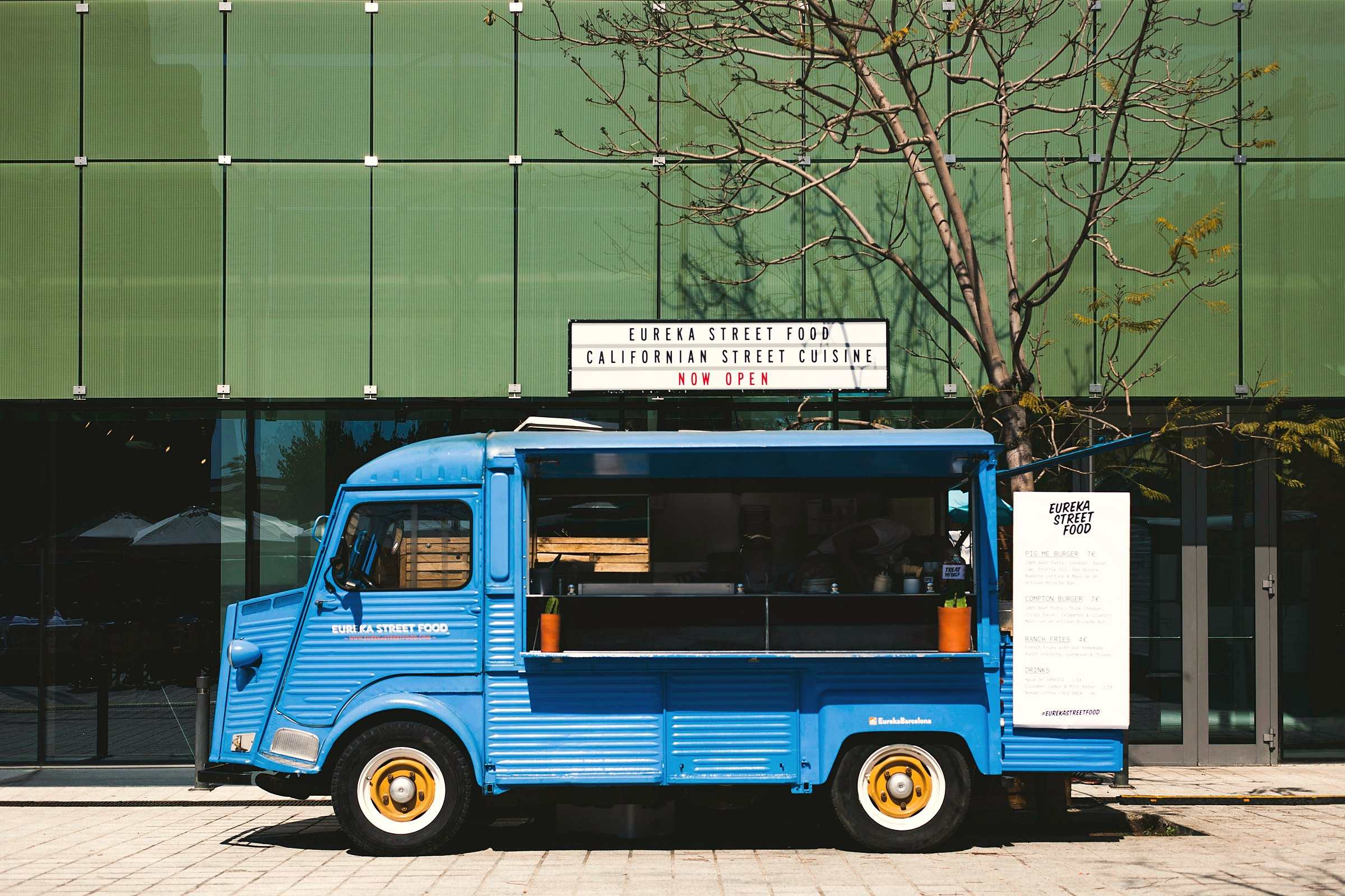 A blue vintage food truck with yellow wheels parked in front of a green glass wall