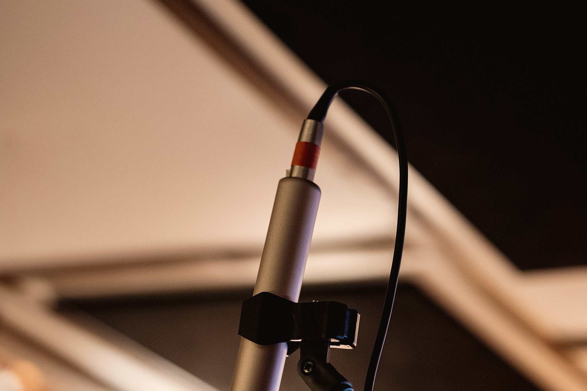 A slim studio microphone on a stand, its cable looping against warm, blurred ceiling beams