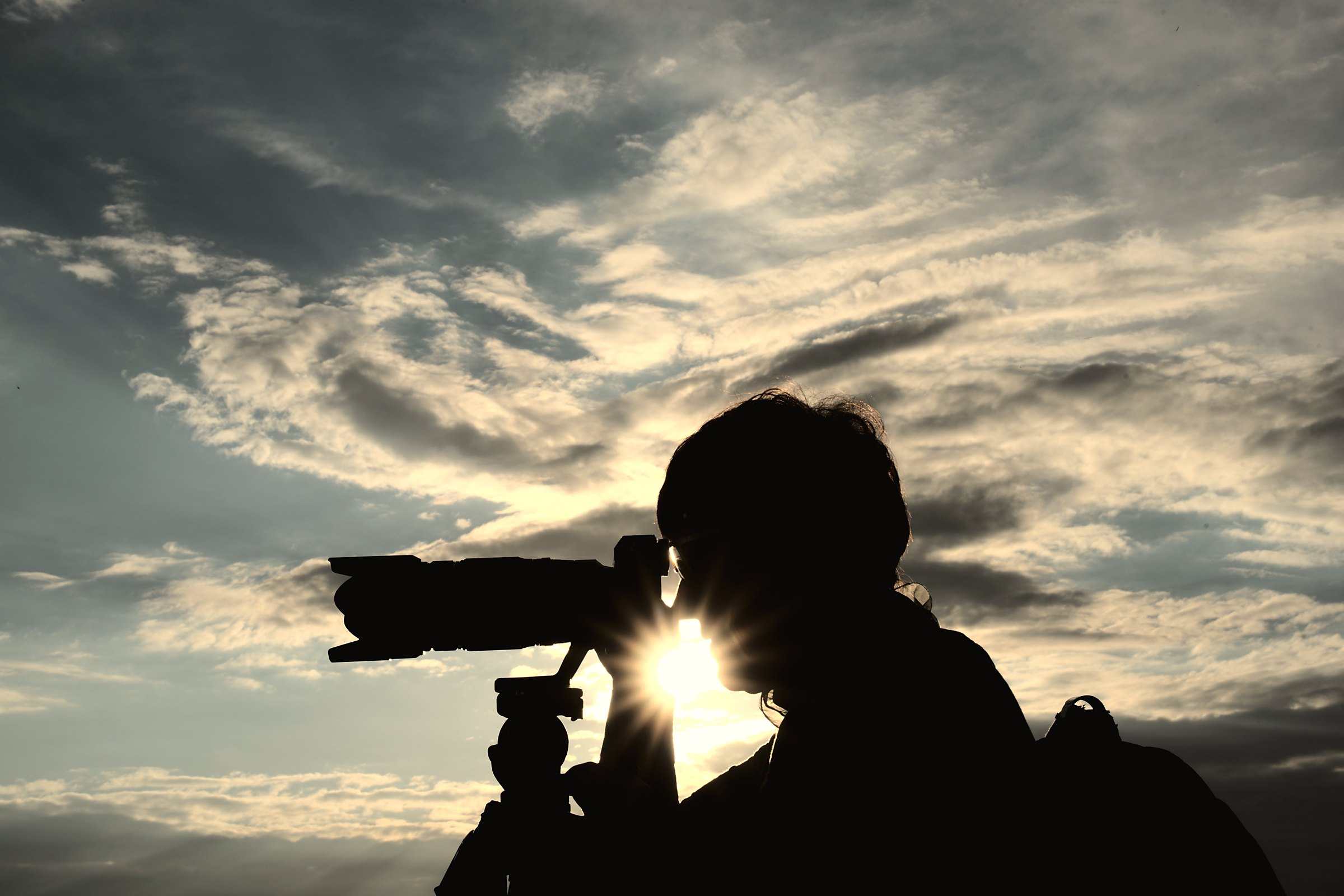 The silhouette of a photographer and a long lens against a bright sunset sky