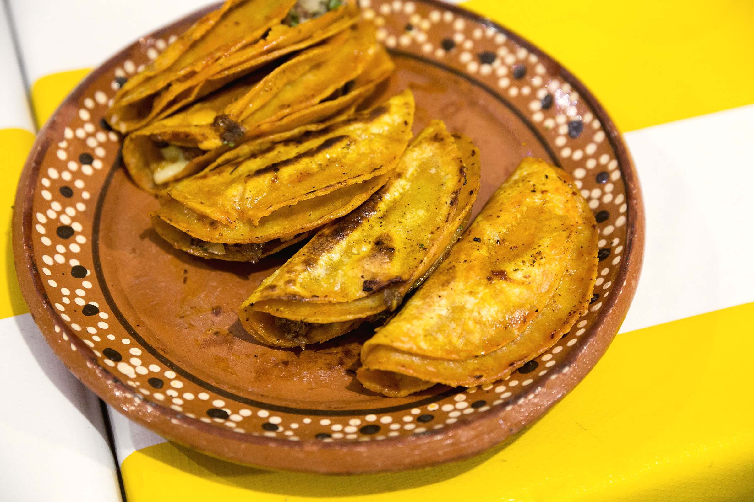 Four folded quesabirria tacos on a hand-painted clay plate over a yellow-striped tablecloth