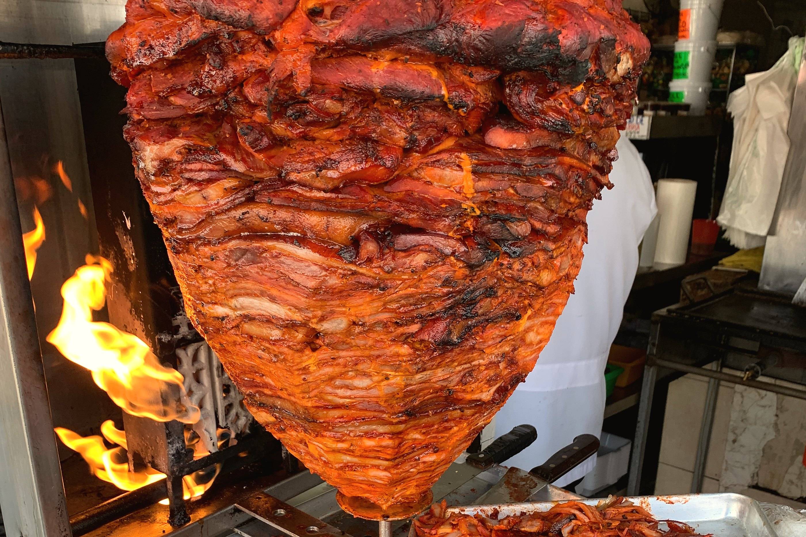 A trompo of al pastor pork turning next to an open flame in a taquería kitchen