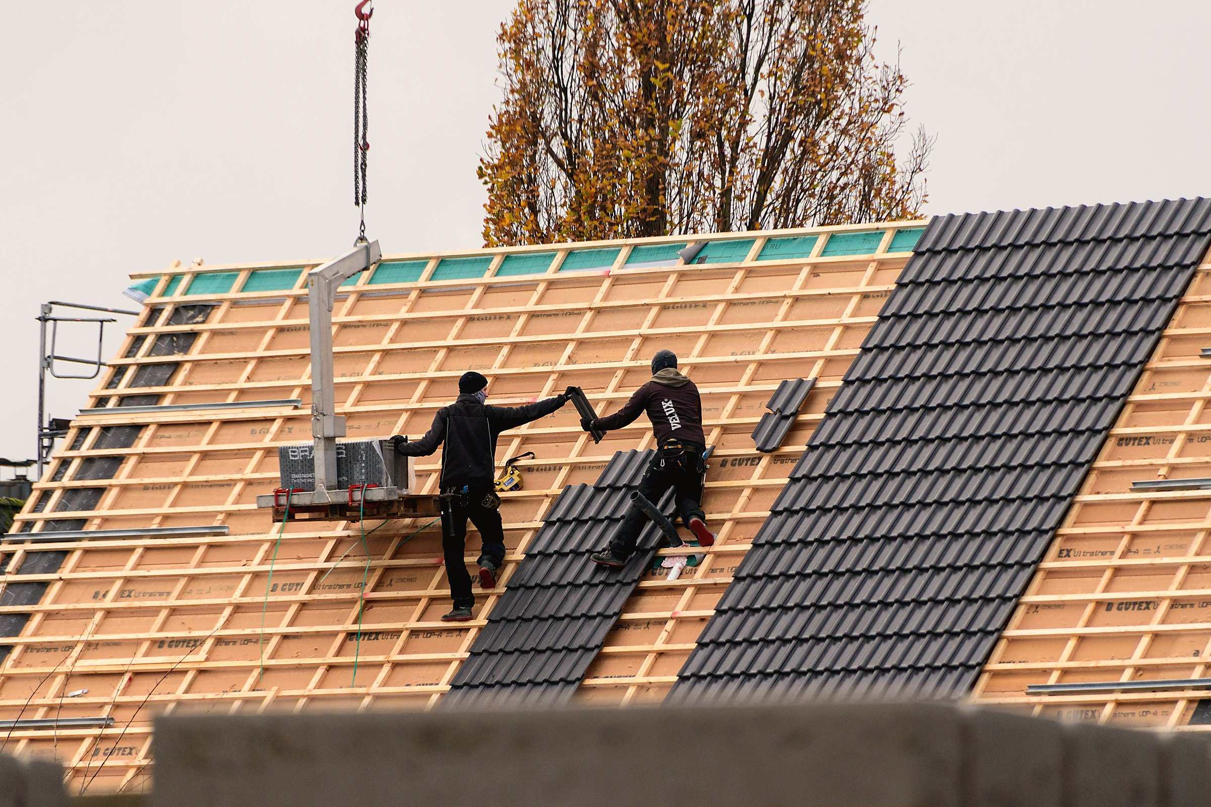 Two roofers laying dark tiles on a new roof while a crane lowers materials