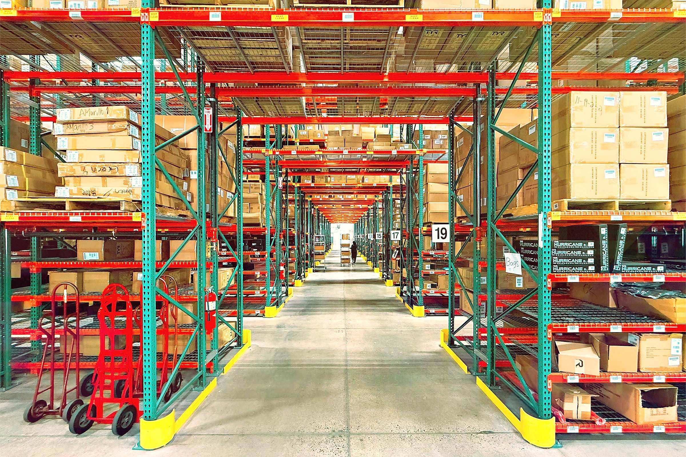 A long warehouse aisle between red and teal pallet racks stacked with boxes