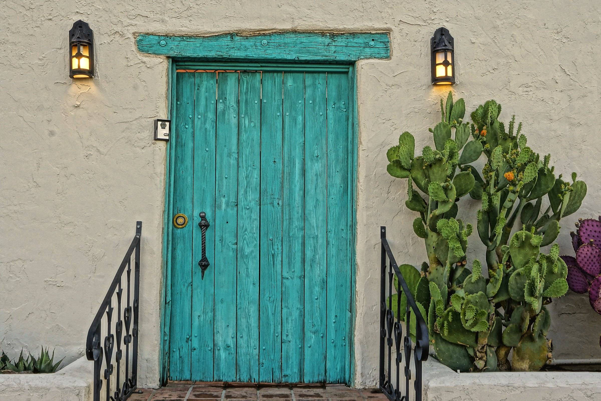 A turquoise wooden door in a white stucco wall in Mesilla, with a prickly pear and two lanterns