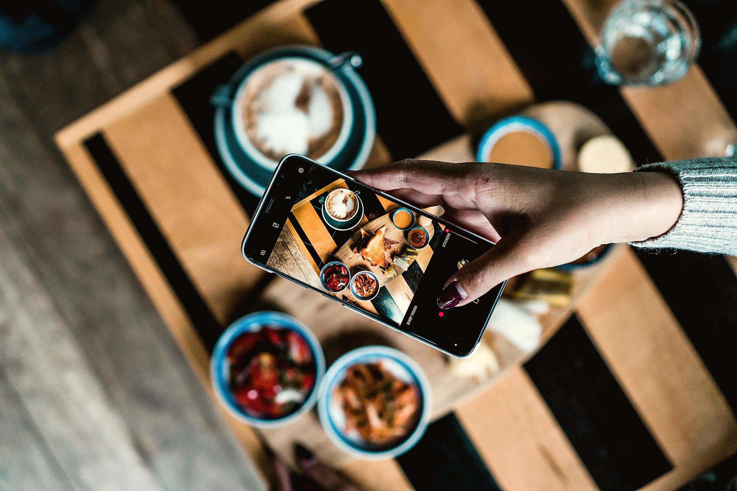 A hand photographing coffee and snacks from above with a smartphone
