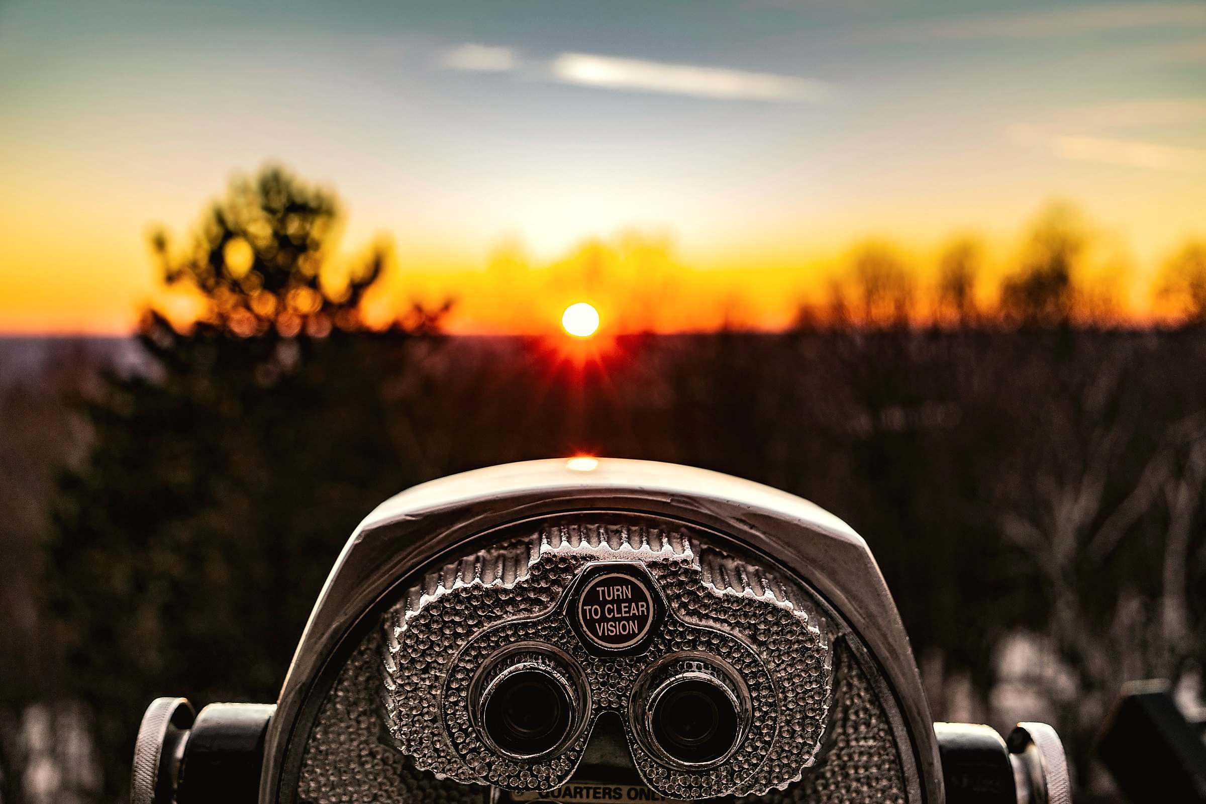 A coin-operated viewing telescope facing a sunset over the trees
