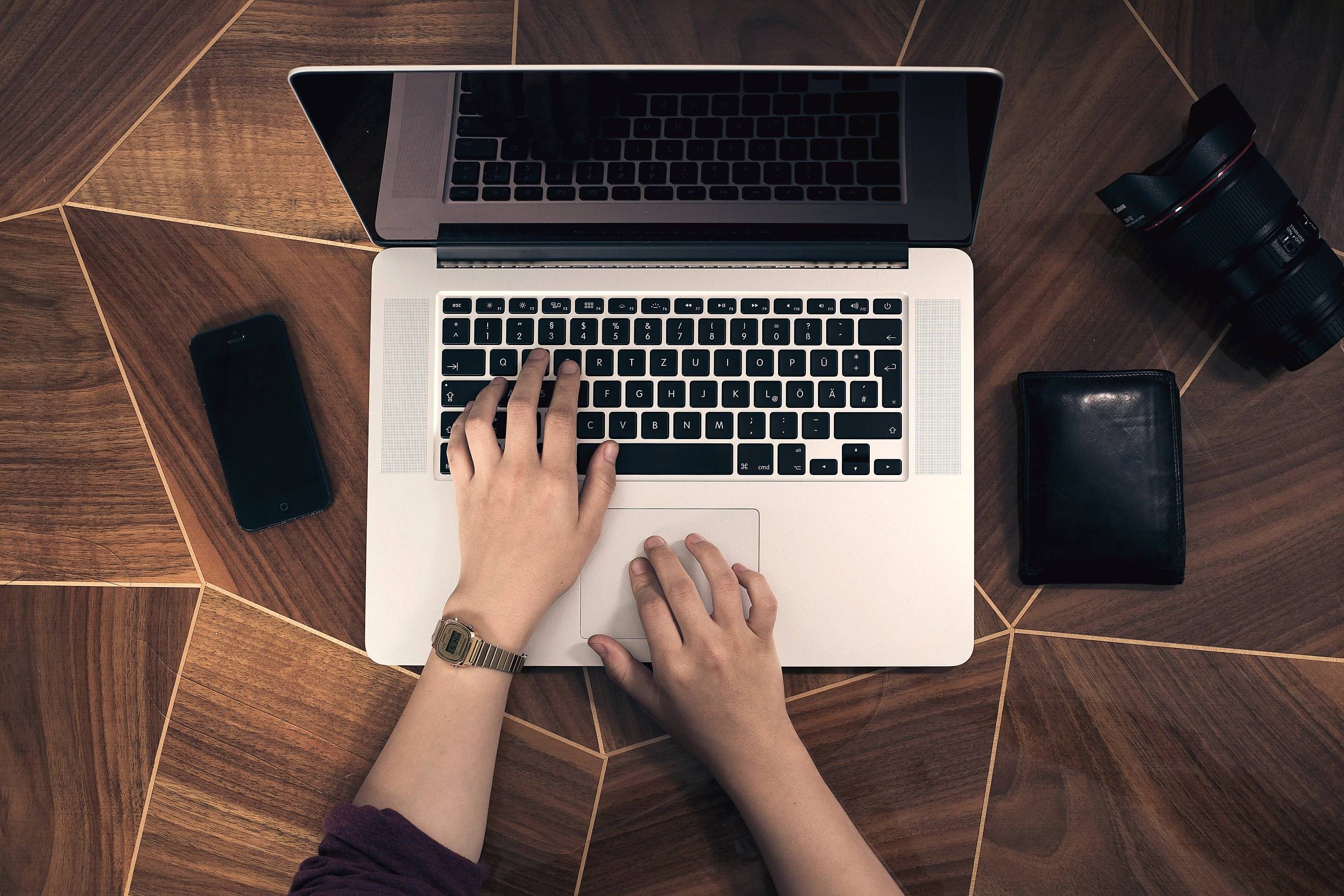 Hands typing on a laptop seen from above, on a patterned walnut floor with a phone, a camera lens and a wallet